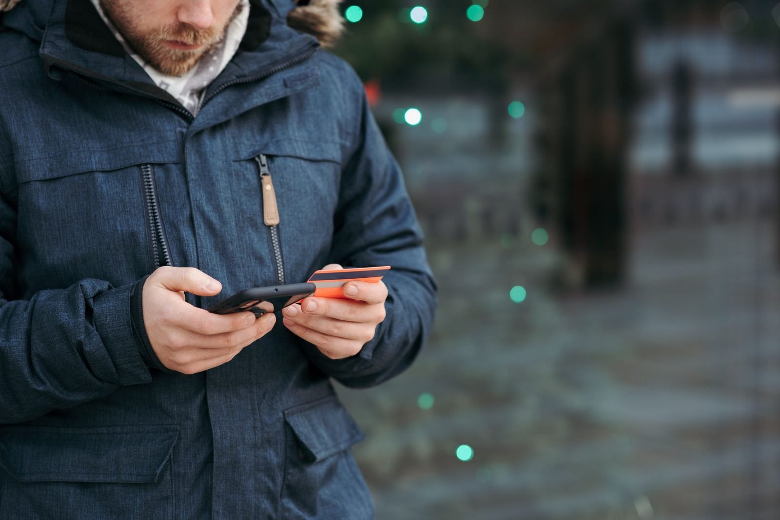 Homem segurando o celular e um cartão bancário numa rua, com luzes desfocadas ao fundo