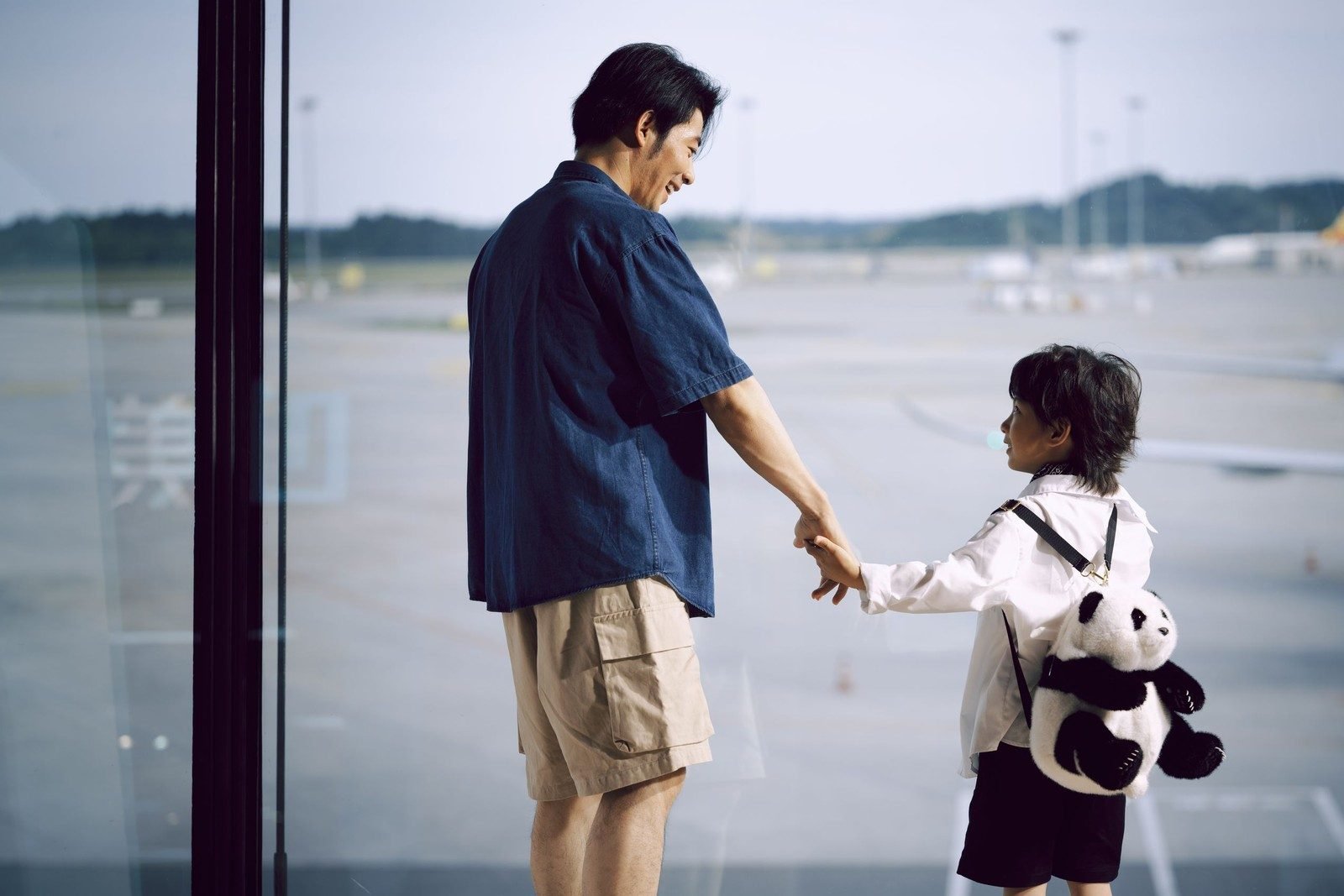 Pai e filho de mãos dadas diante da janela de um aeroporto, com a pista ao fundo